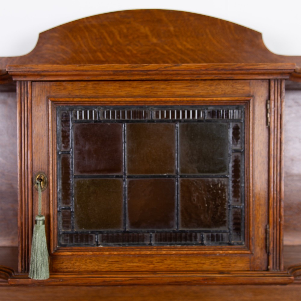An Arts and Crafts oak cabinet dresser featuring an upper shelf unit with a central leaded glass door above a base section with a single drawer, cupboard doors, and an open side alcove. View VII