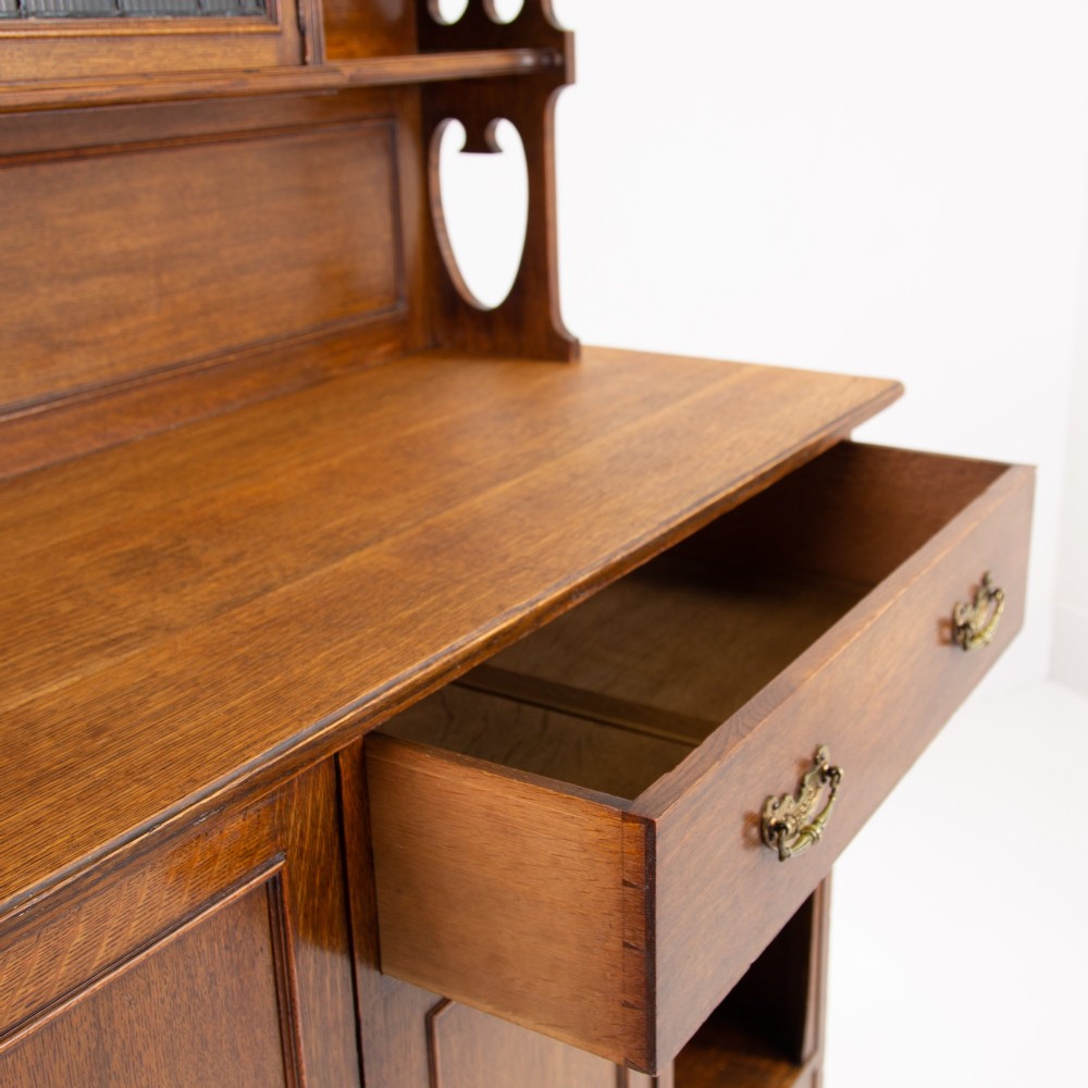 An Arts and Crafts oak cabinet dresser featuring an upper shelf unit with a central leaded glass door above a base section with a single drawer, cupboard doors, and an open side alcove. View VIII
