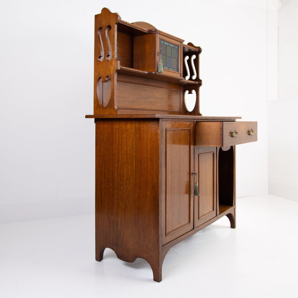 An Arts and Crafts oak cabinet dresser featuring an upper shelf unit with a central leaded glass door above a base section with a single drawer, cupboard doors, and an open side alcove. View IX