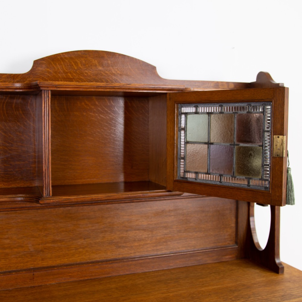 An Arts and Crafts oak cabinet dresser featuring an upper shelf unit with a central leaded glass door above a base section with a single drawer, cupboard doors, and an open side alcove. View XII