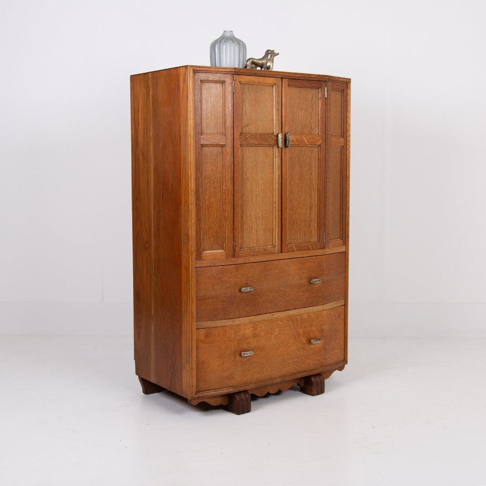 A 1930s Art Deco oak tallboy cabinet featuring a two-door upper cupboard with geometric paneling above two wide drawers, resting on stylized stepped feet.