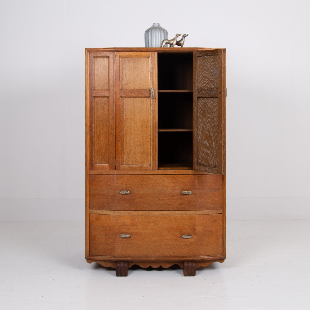 A 1930s Art Deco oak tallboy cabinet featuring a two-door upper cupboard with geometric paneling above two wide drawers, resting on stylized stepped feet. View IV