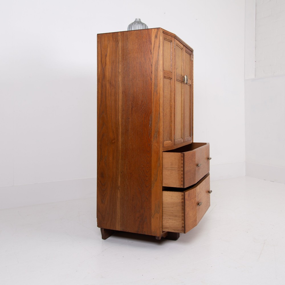 A 1930s Art Deco oak tallboy cabinet featuring a two-door upper cupboard with geometric paneling above two wide drawers, resting on stylized stepped feet. View V
