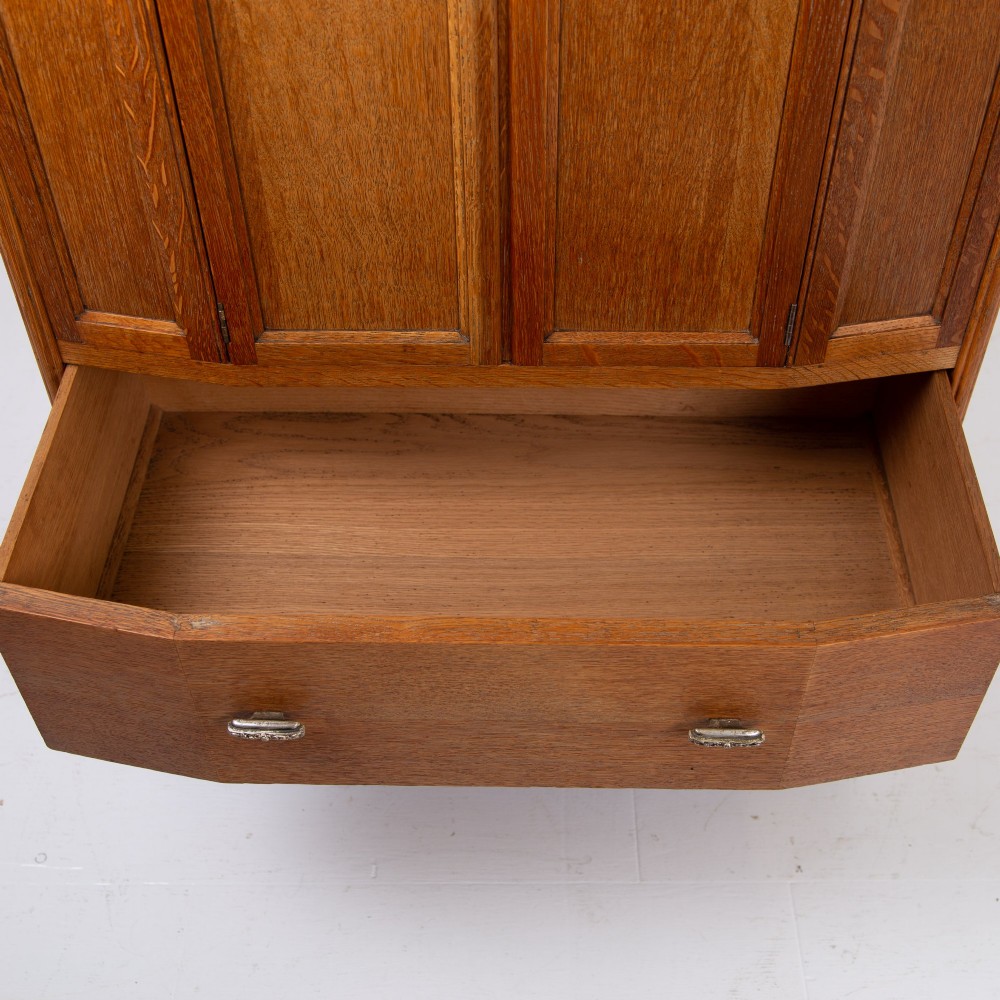 A 1930s Art Deco oak tallboy cabinet featuring a two-door upper cupboard with geometric paneling above two wide drawers, resting on stylized stepped feet. View VI