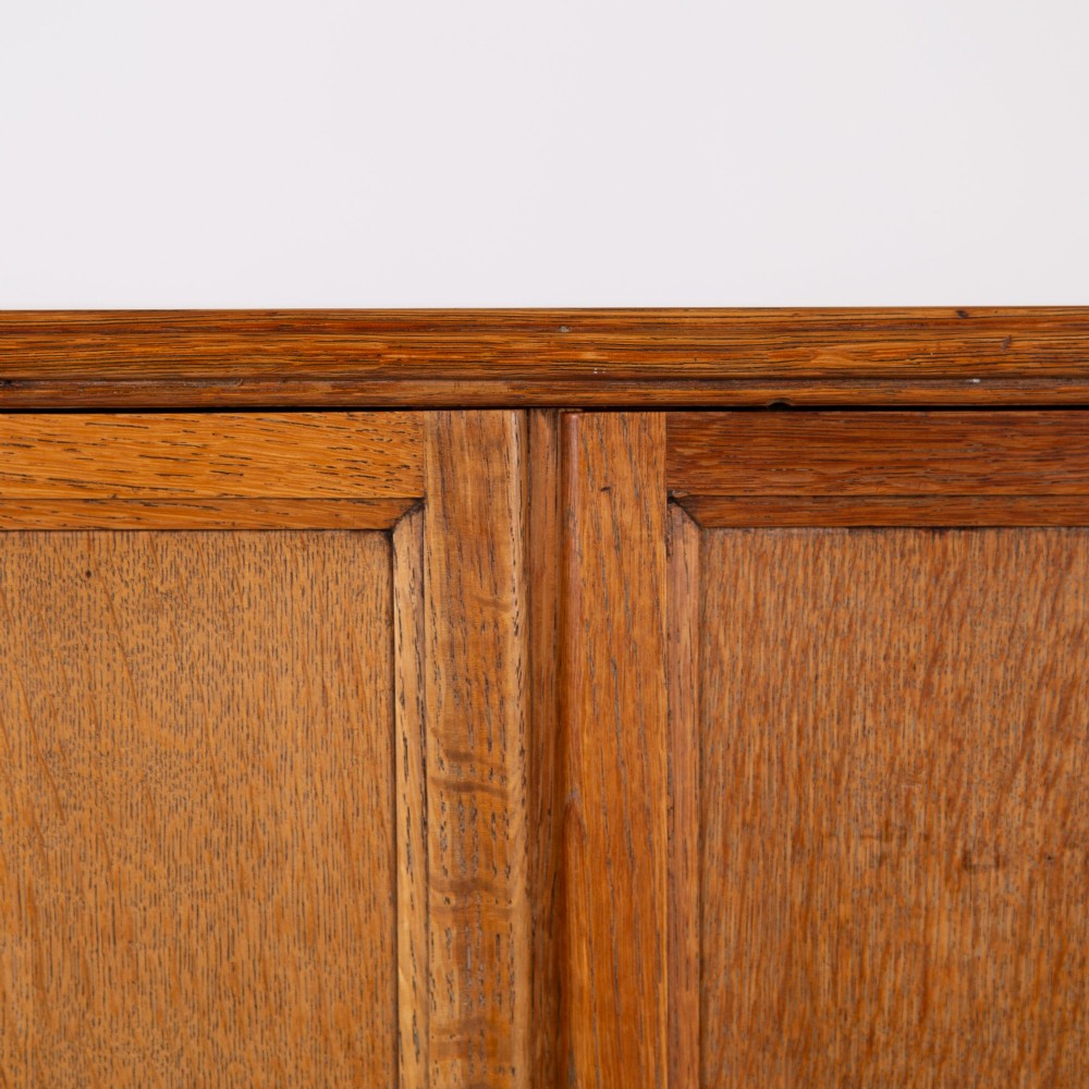 A 1930s Art Deco oak tallboy cabinet featuring a two-door upper cupboard with geometric paneling above two wide drawers, resting on stylized stepped feet. View VII
