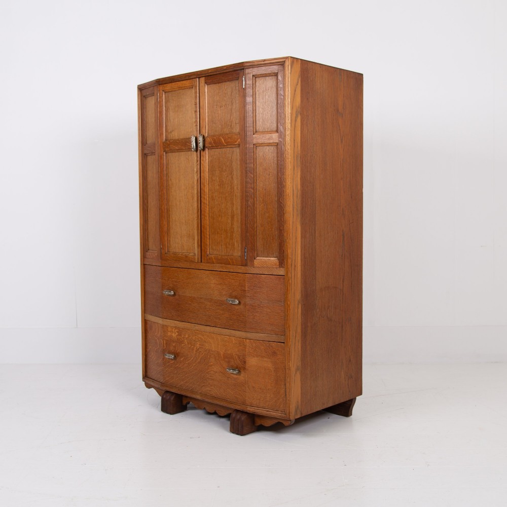 A 1930s Art Deco oak tallboy cabinet featuring a two-door upper cupboard with geometric paneling above two wide drawers, resting on stylized stepped feet. View XIV