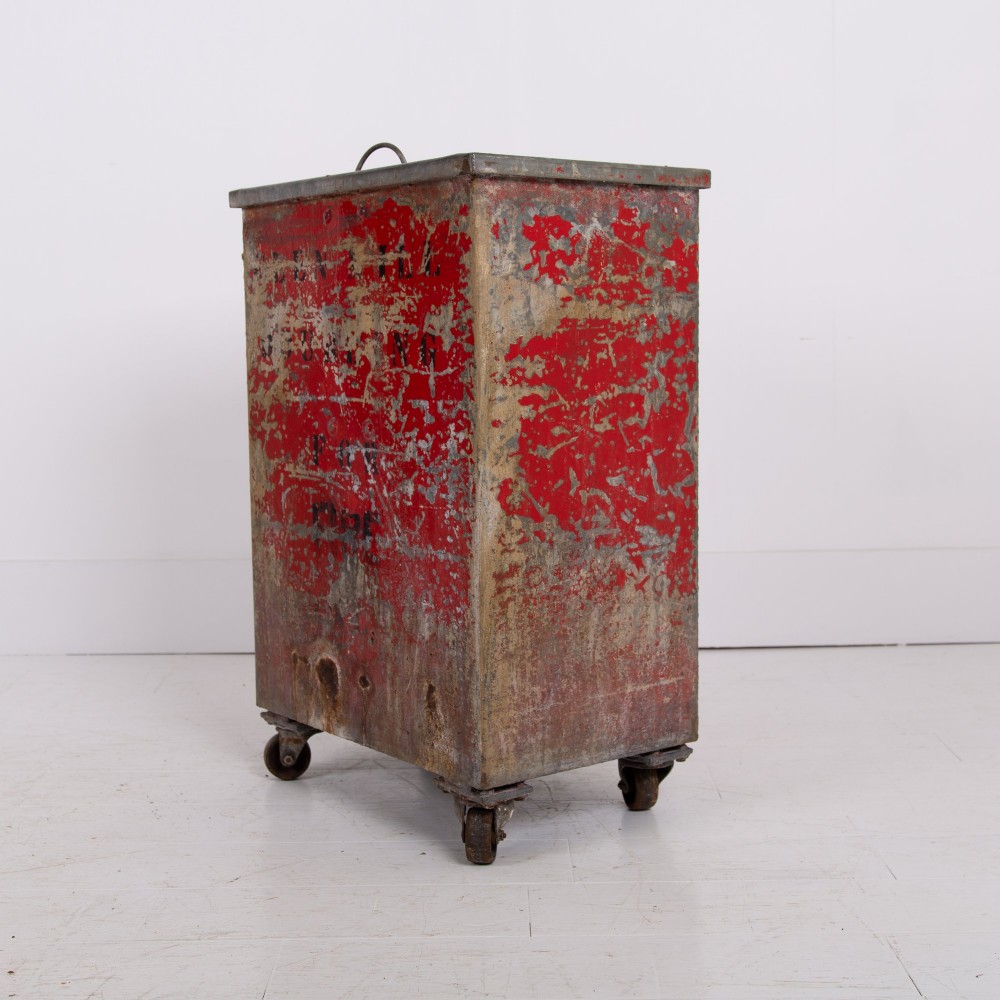 A tall rectangular industrial metal fire bin with distressed red paint, stencilled black text, a top lid, and four small cast iron swivel castors.