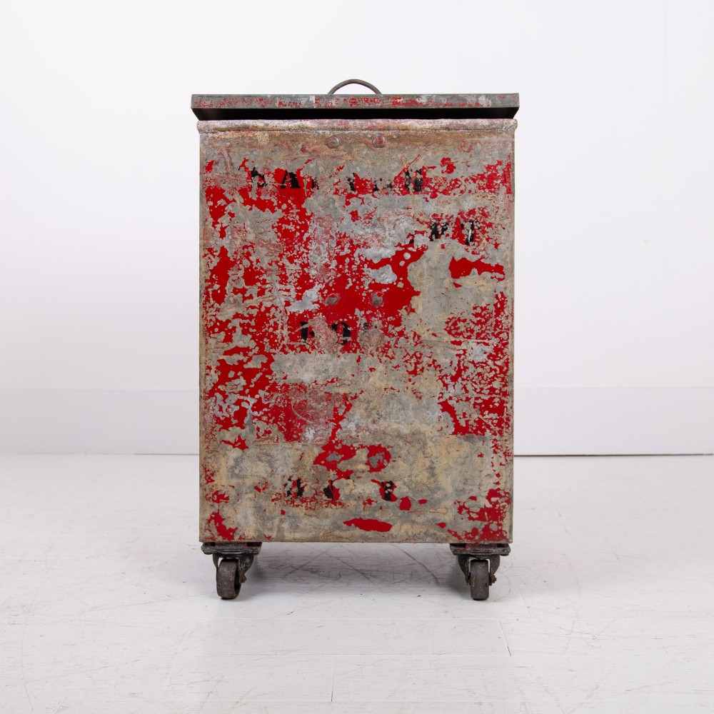 A tall rectangular industrial metal fire bin with distressed red paint, stencilled black text, a top lid, and four small cast iron swivel castors. View II
