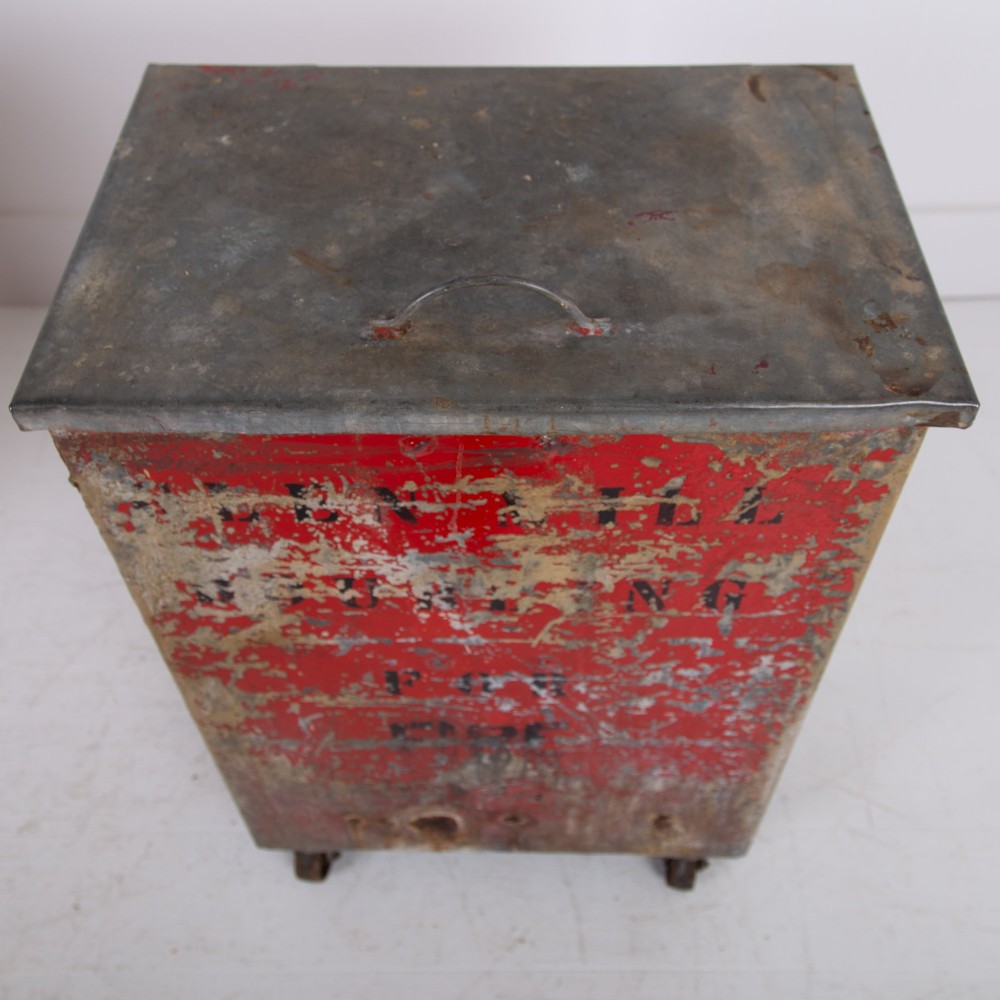 A tall rectangular industrial metal fire bin with distressed red paint, stencilled black text, a top lid, and four small cast iron swivel castors. View IV