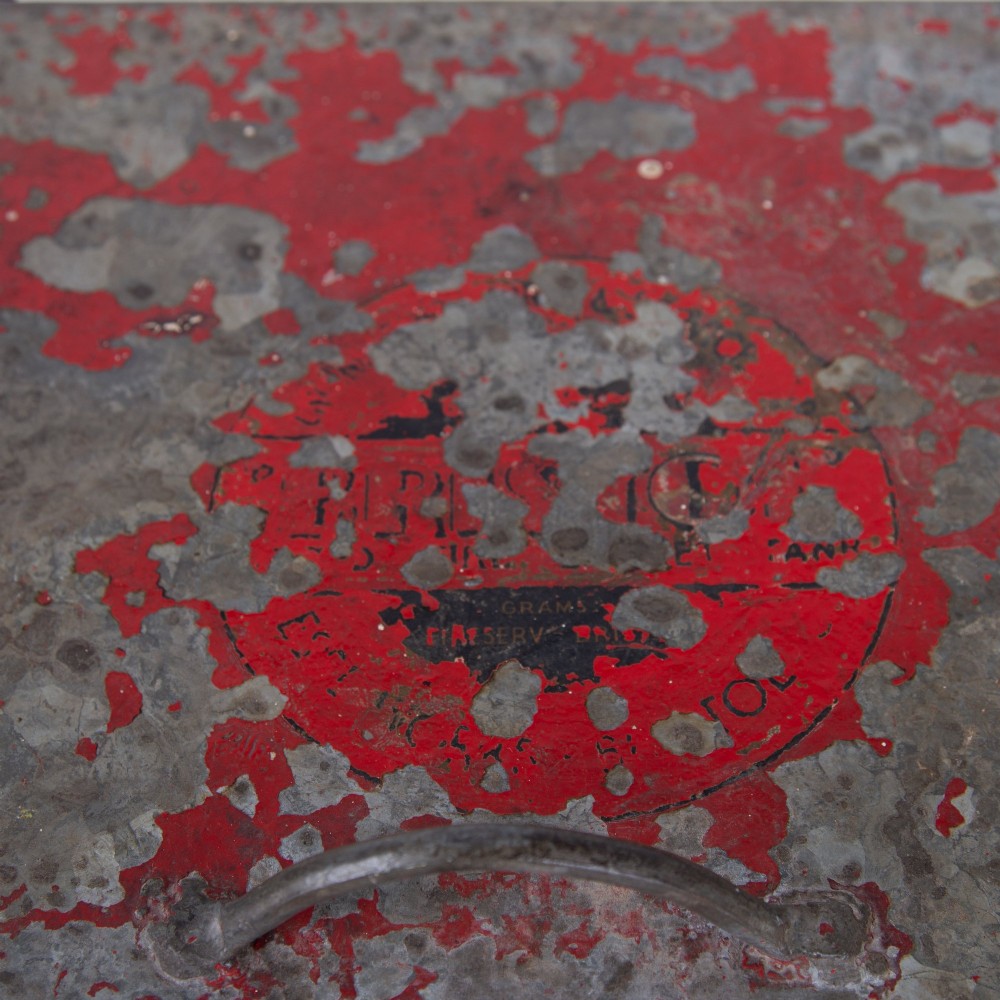 A tall rectangular industrial metal fire bin with distressed red paint, stencilled black text, a top lid, and four small cast iron swivel castors. View V