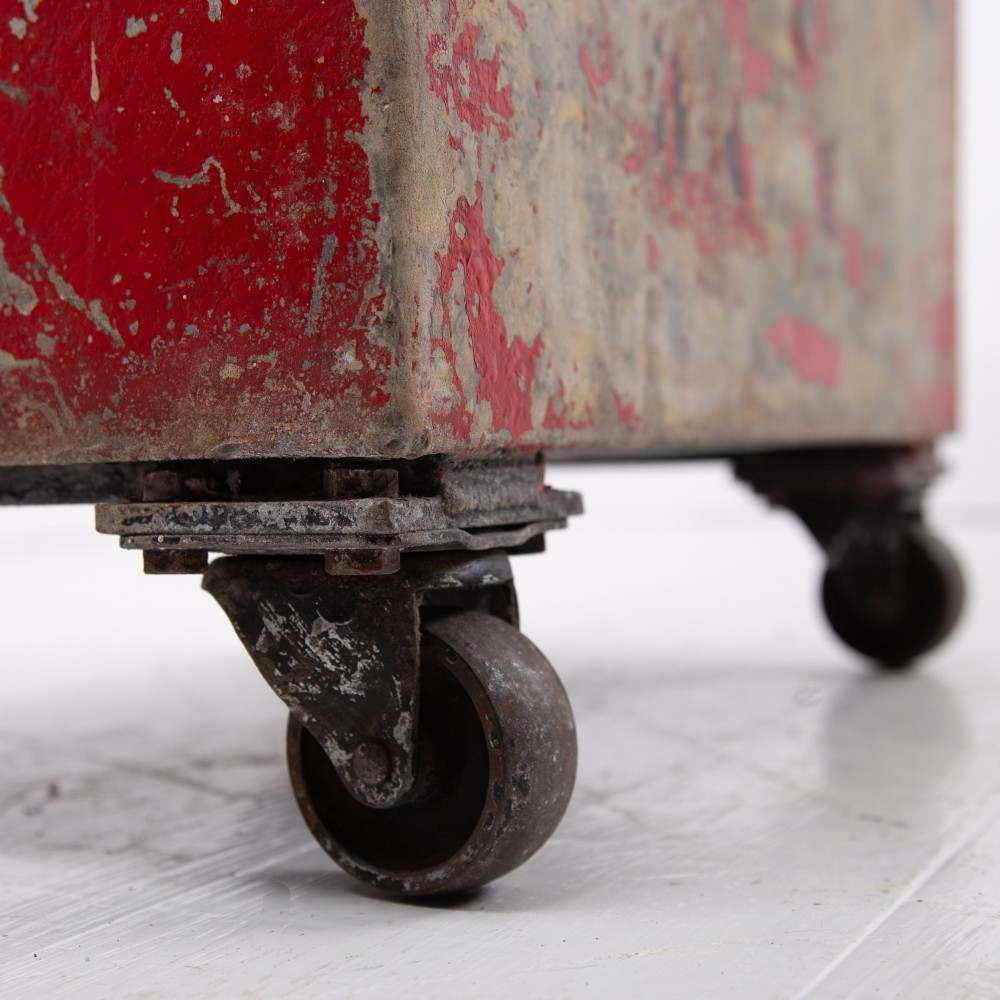 A tall rectangular industrial metal fire bin with distressed red paint, stencilled black text, a top lid, and four small cast iron swivel castors. View VI