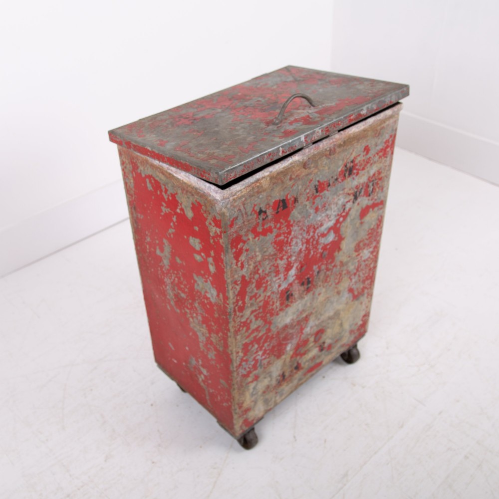 A tall rectangular industrial metal fire bin with distressed red paint, stencilled black text, a top lid, and four small cast iron swivel castors. View VII