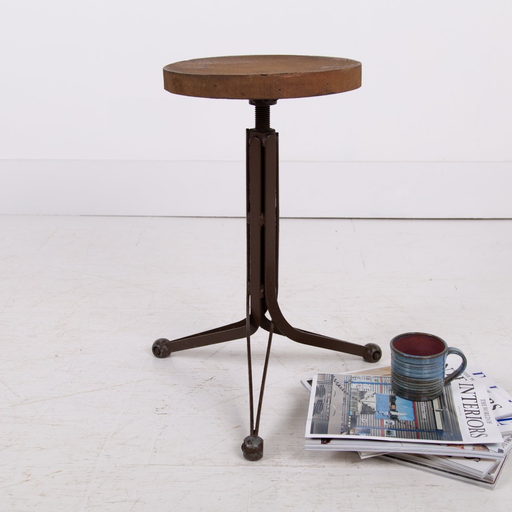 An industrial 1930s workshop stool featuring a circular wooden seat mounted on a dark brown metal tripod base with a threaded height adjustment mechanism.