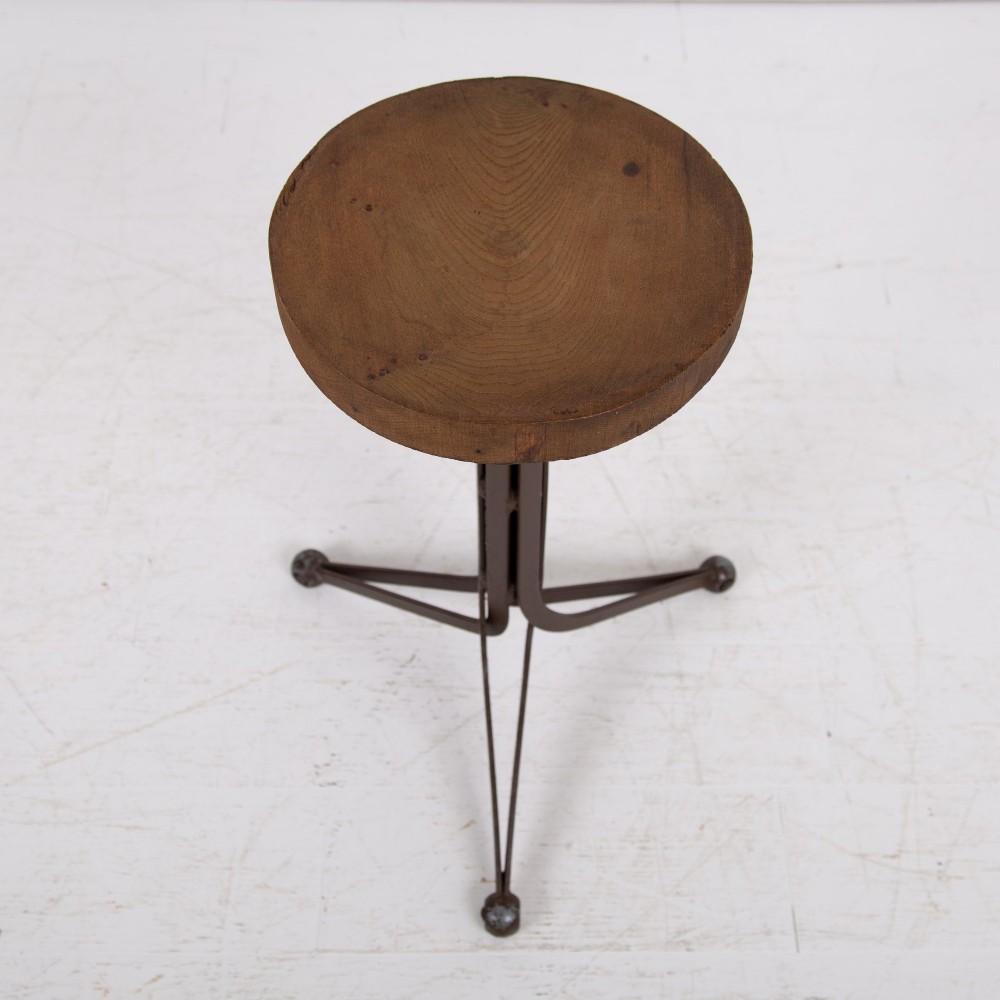 An industrial 1930s workshop stool featuring a circular wooden seat mounted on a dark brown metal tripod base with a threaded height adjustment mechanism. View II