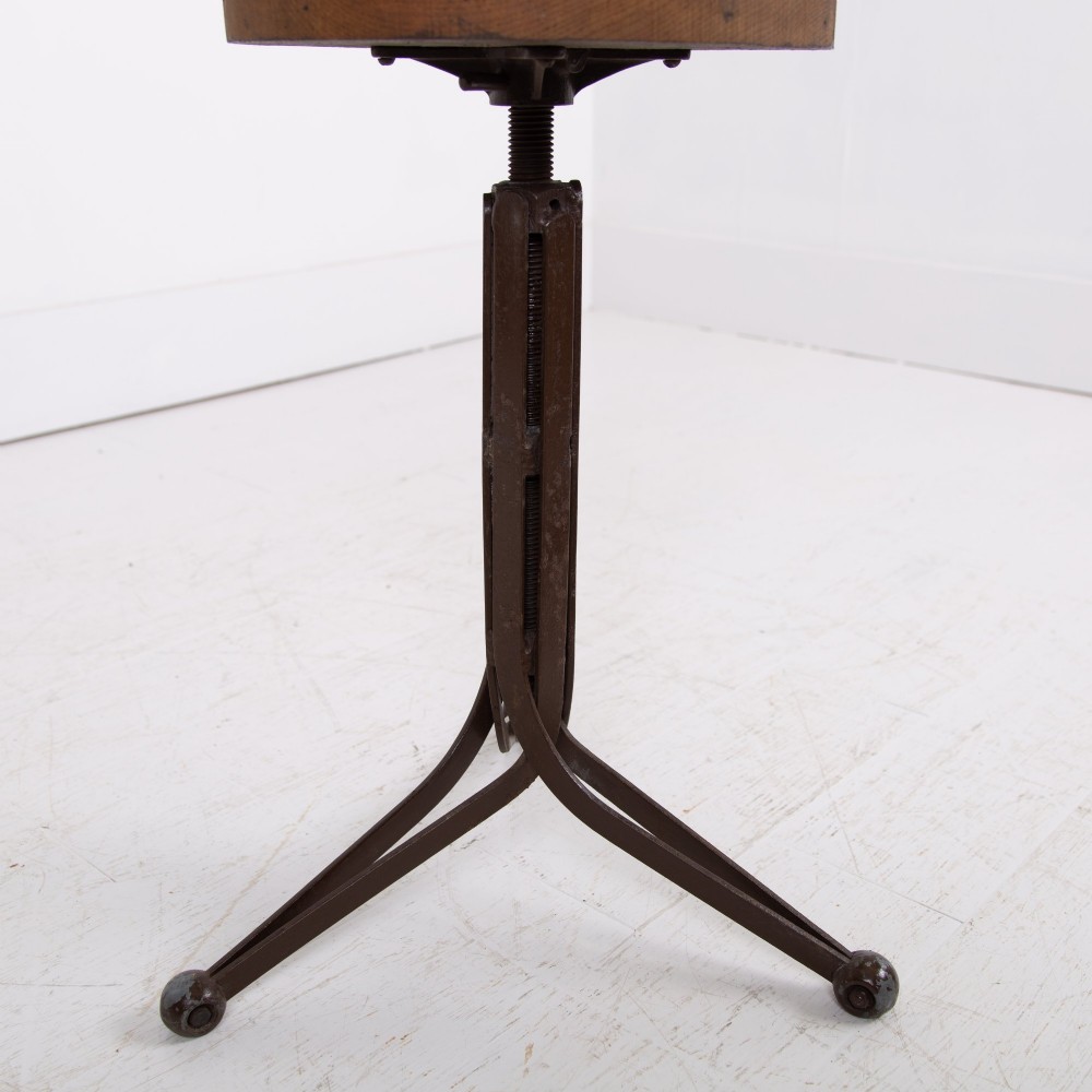 An industrial 1930s workshop stool featuring a circular wooden seat mounted on a dark brown metal tripod base with a threaded height adjustment mechanism. View VII