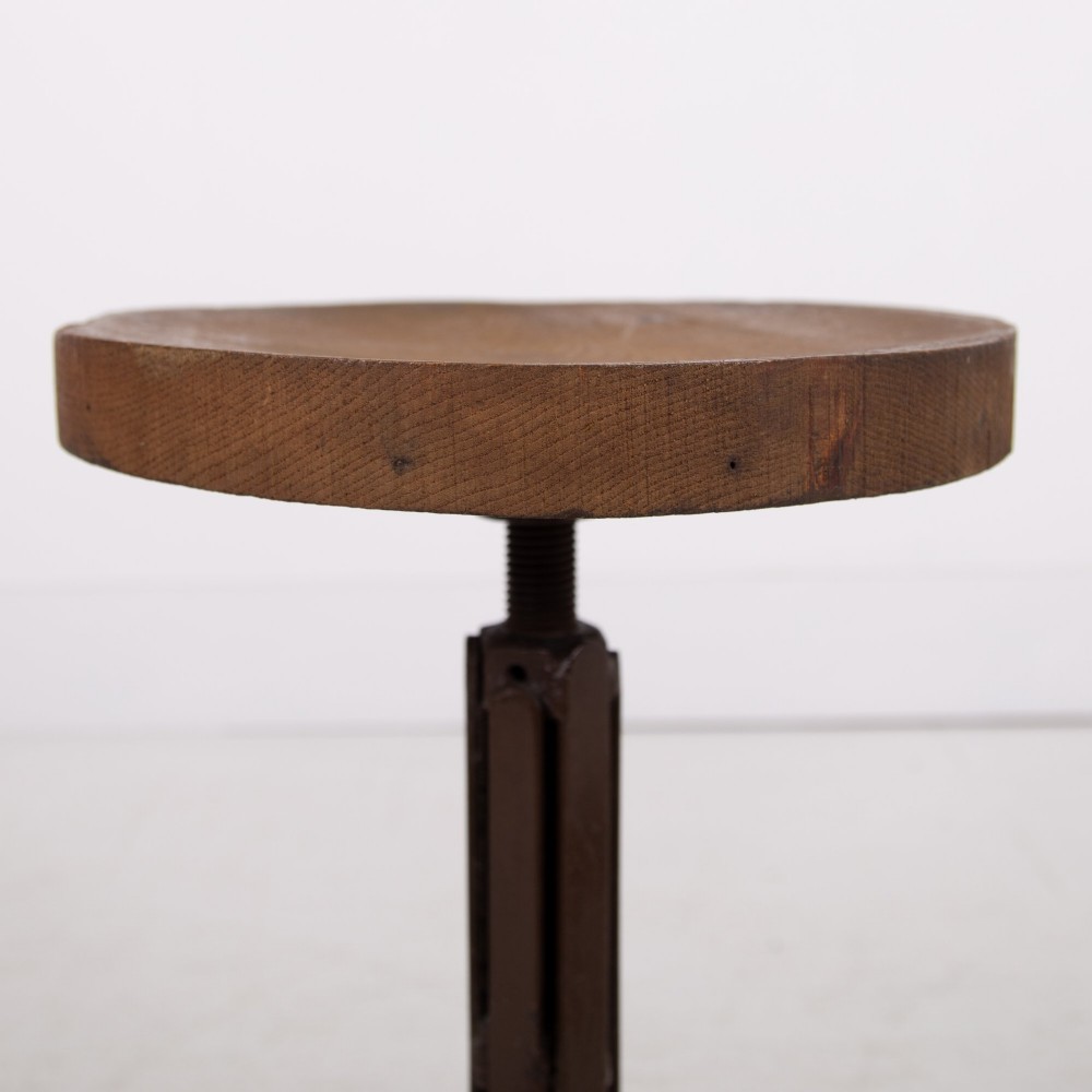 An industrial 1930s workshop stool featuring a circular wooden seat mounted on a dark brown metal tripod base with a threaded height adjustment mechanism. View IX