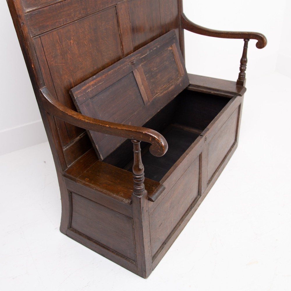 A 19th-century dark oak high-back box settle with panelled detailing, scrolled armrests, turned supports, and a hinged seat opening to a box storage base. View IV