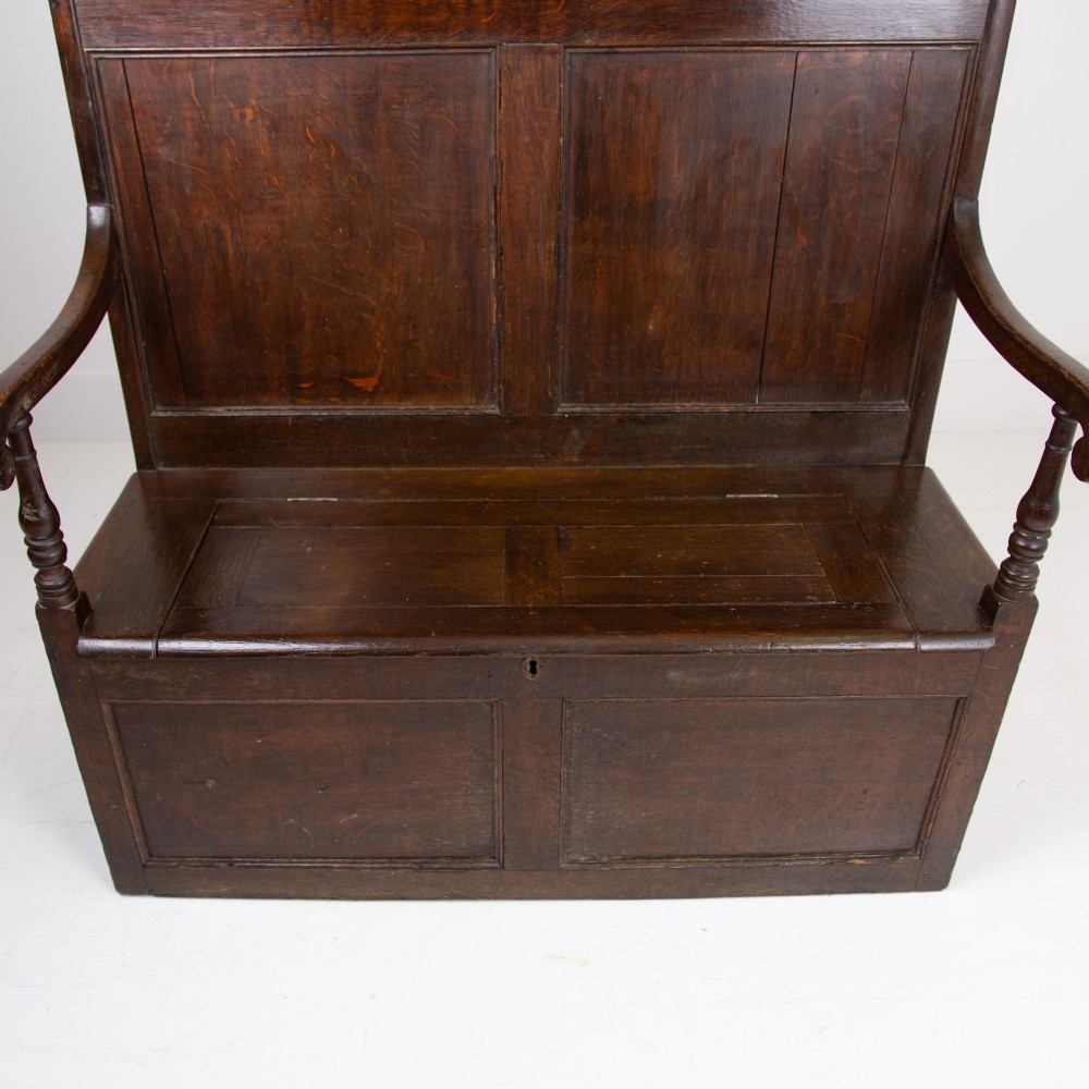 A 19th-century dark oak high-back box settle with panelled detailing, scrolled armrests, turned supports, and a hinged seat opening to a box storage base. View V