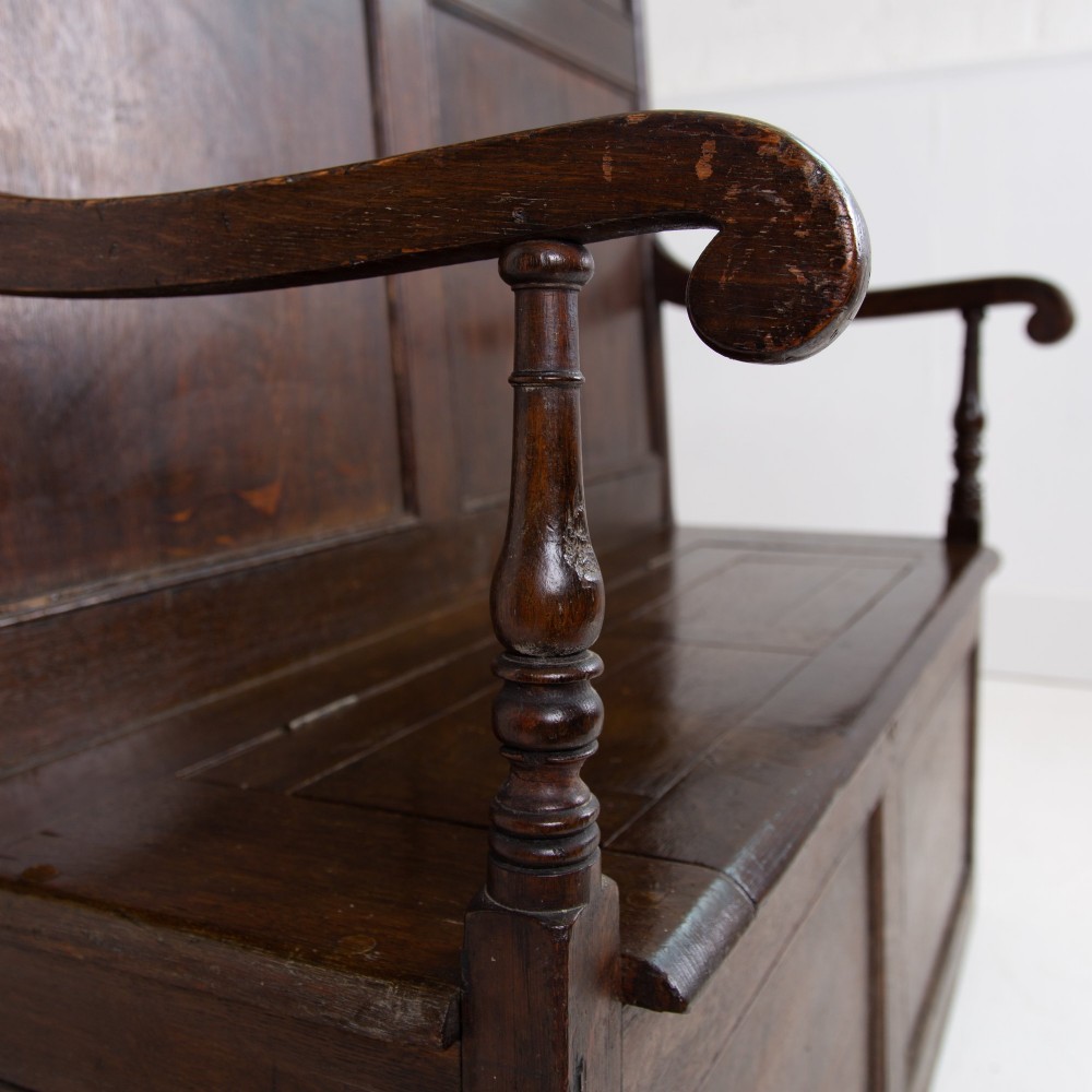 A 19th-century dark oak high-back box settle with panelled detailing, scrolled armrests, turned supports, and a hinged seat opening to a box storage base. View VI