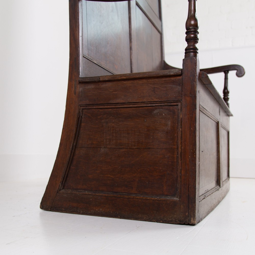 A 19th-century dark oak high-back box settle with panelled detailing, scrolled armrests, turned supports, and a hinged seat opening to a box storage base. View VII