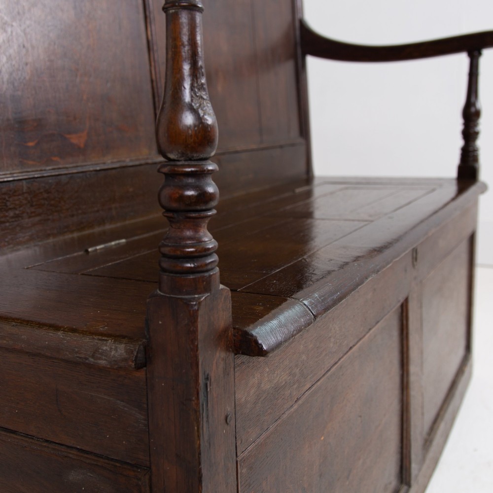A 19th-century dark oak high-back box settle with panelled detailing, scrolled armrests, turned supports, and a hinged seat opening to a box storage base. View VIII