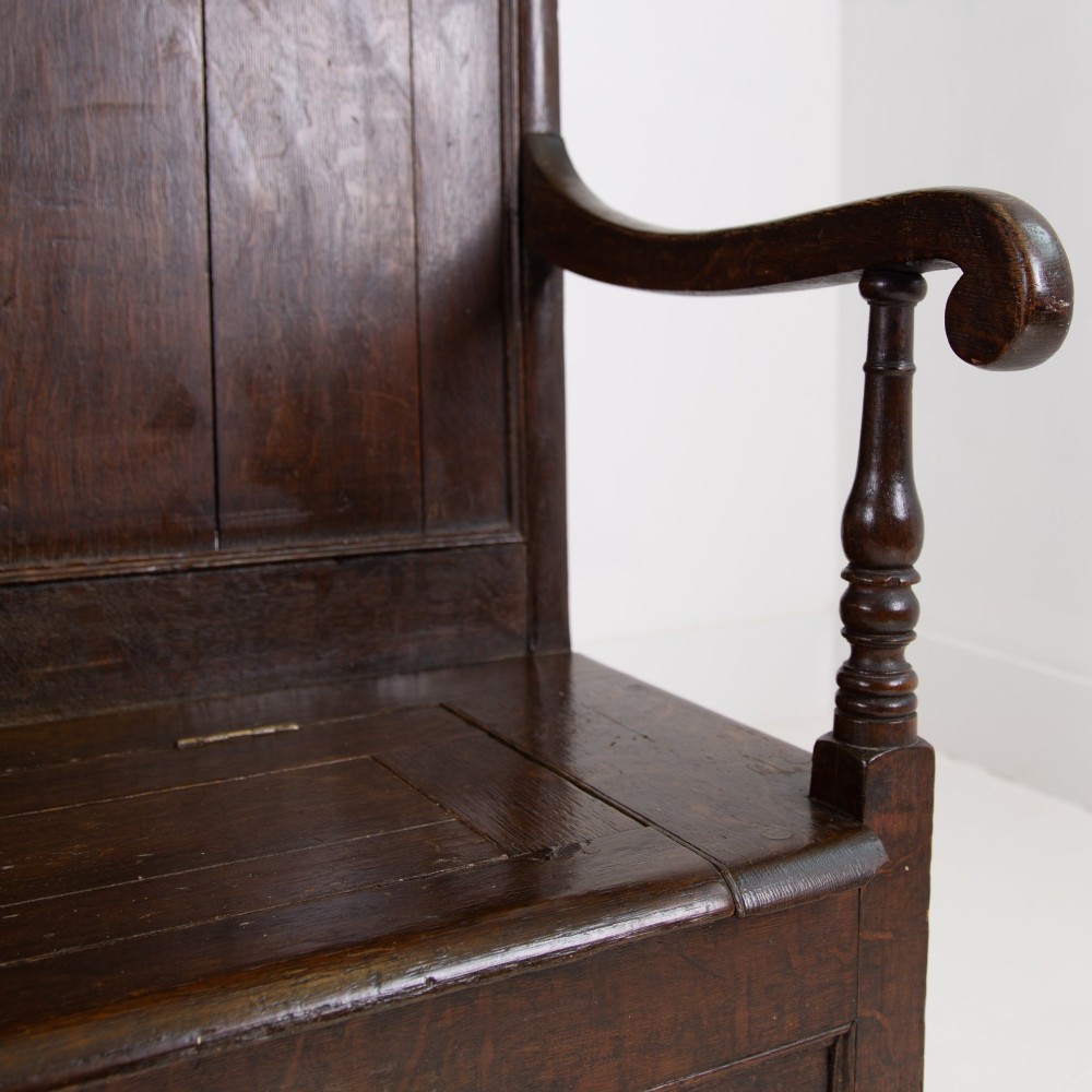 A 19th-century dark oak high-back box settle with panelled detailing, scrolled armrests, turned supports, and a hinged seat opening to a box storage base. View IX