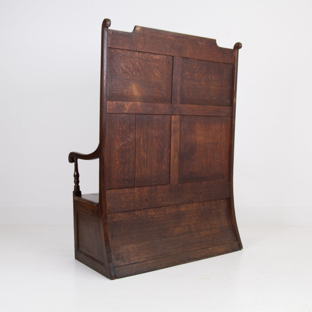 A 19th-century dark oak high-back box settle with panelled detailing, scrolled armrests, turned supports, and a hinged seat opening to a box storage base. View XI