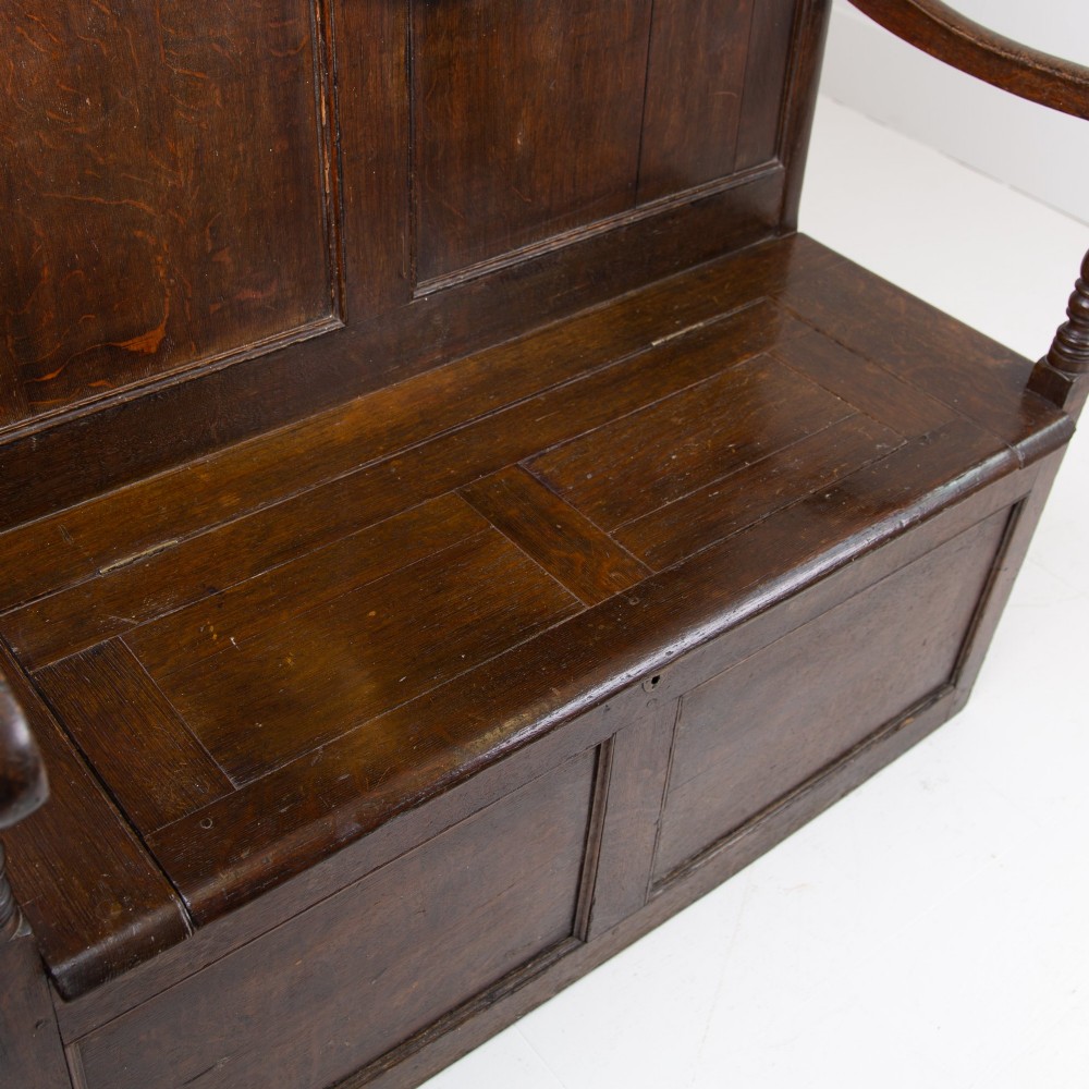A 19th-century dark oak high-back box settle with panelled detailing, scrolled armrests, turned supports, and a hinged seat opening to a box storage base. View XII