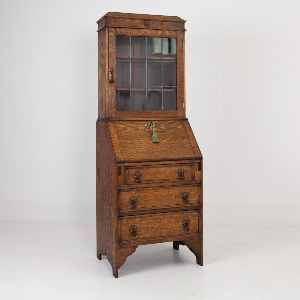 A small early 20th-century oak bureau bookcase featuring a leaded glass upper cabinet, a slant-front writing slope, and three lower drawers with brass drop handles.