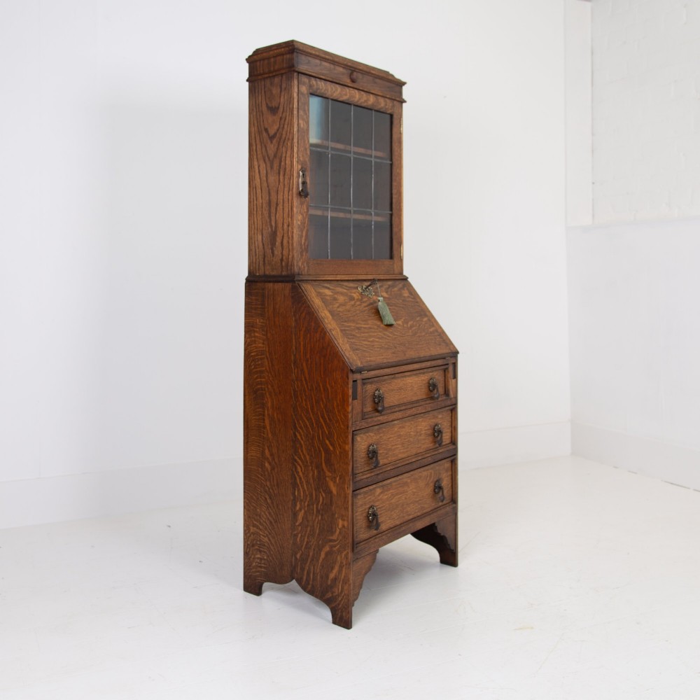 A small early 20th-century oak bureau bookcase featuring a leaded glass upper cabinet, a slant-front writing slope, and three lower drawers with brass drop handles. View II