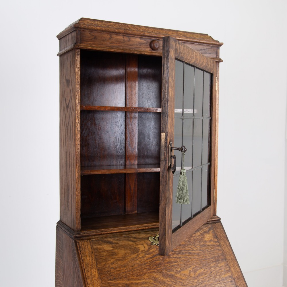 A small early 20th-century oak bureau bookcase featuring a leaded glass upper cabinet, a slant-front writing slope, and three lower drawers with brass drop handles. View VI