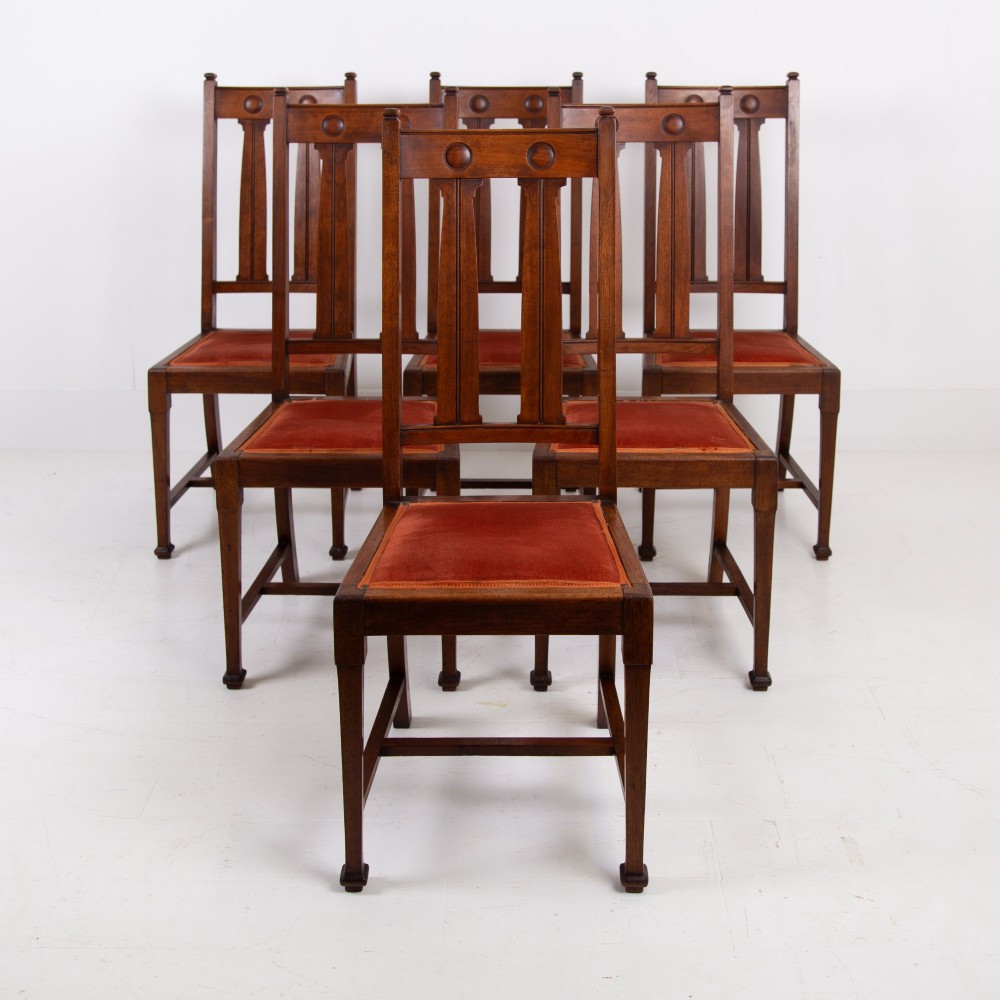 A set of six Scottish Arts and Crafts oak dining chairs featuring high slatted backs with carved roundel details and red velvet seat pads.