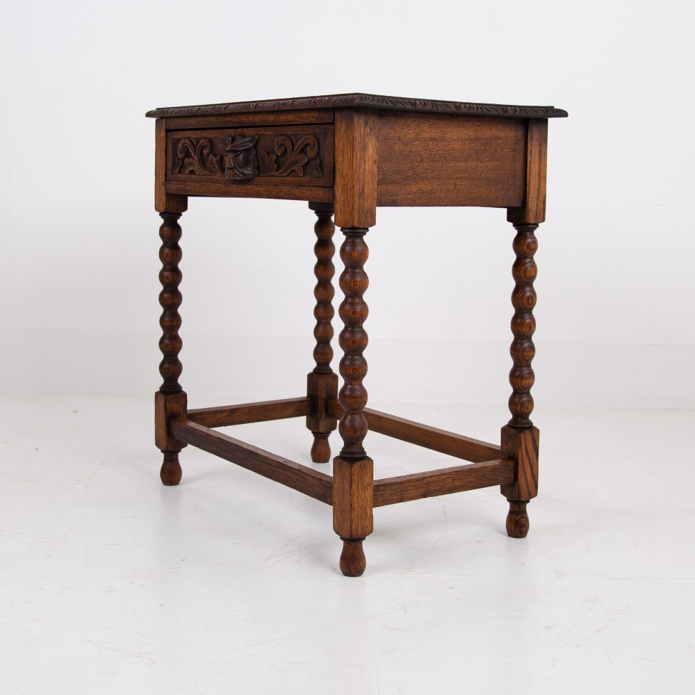 A dark oak Victorian side table featuring a single drawer carved with a Green Man mask motif, bobbin-turned legs, and a lower rectangular stretcher. View X