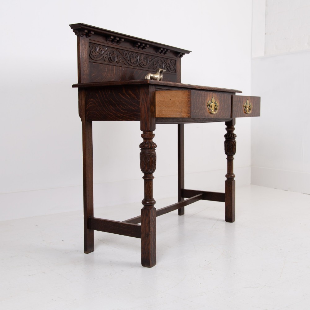 A dark oak console hall table featuring a carved back gallery, two drawers with brass backplate handles, and turned front legs. View X