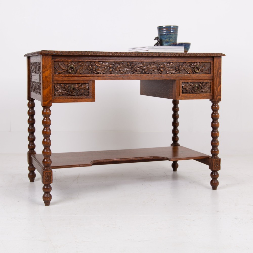 An oak writing desk featuring three carved foliage drawer fronts, bobbin-turned legs, brass drop handles, and a low under-tier shelf.