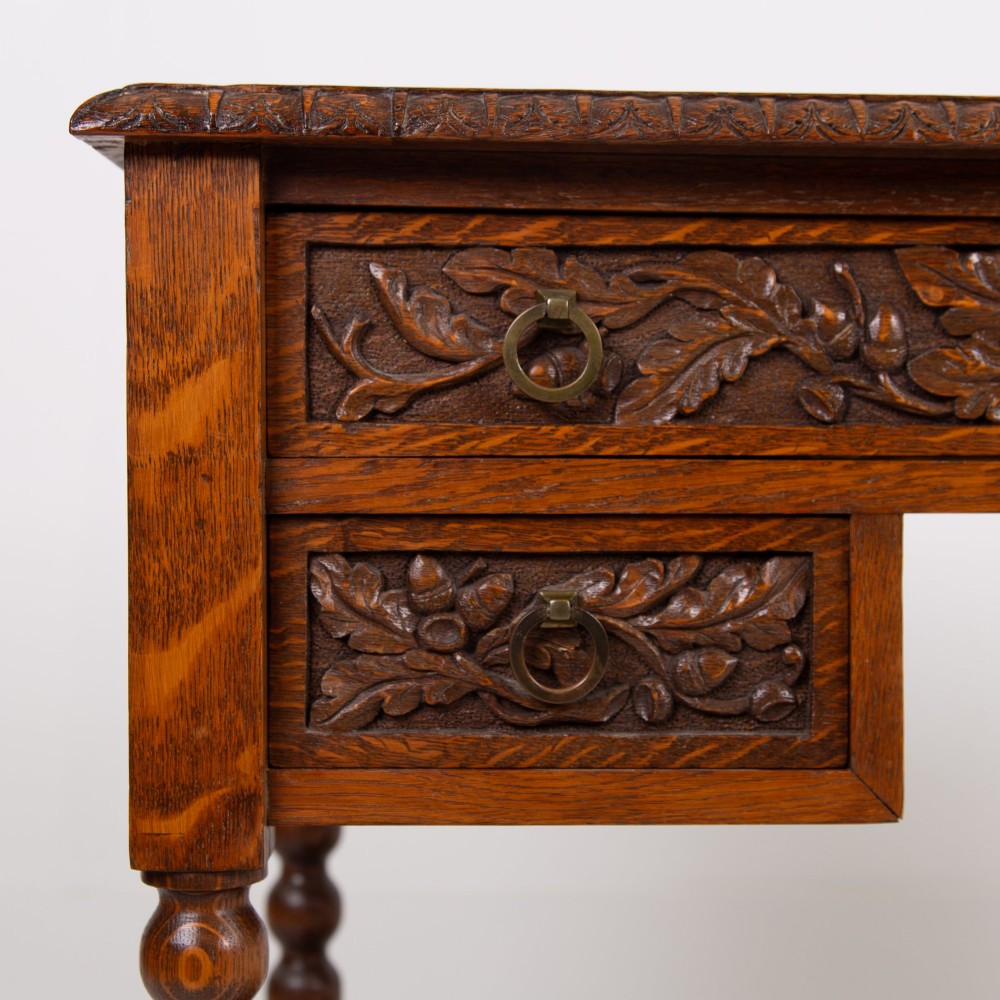 An oak writing desk featuring three carved foliage drawer fronts, bobbin-turned legs, brass drop handles, and a low under-tier shelf. View II