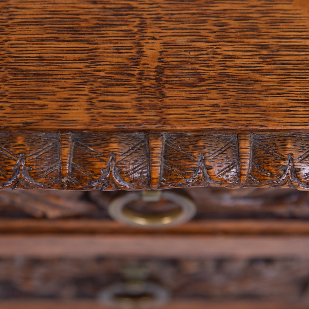 An oak writing desk featuring three carved foliage drawer fronts, bobbin-turned legs, brass drop handles, and a low under-tier shelf. View VII