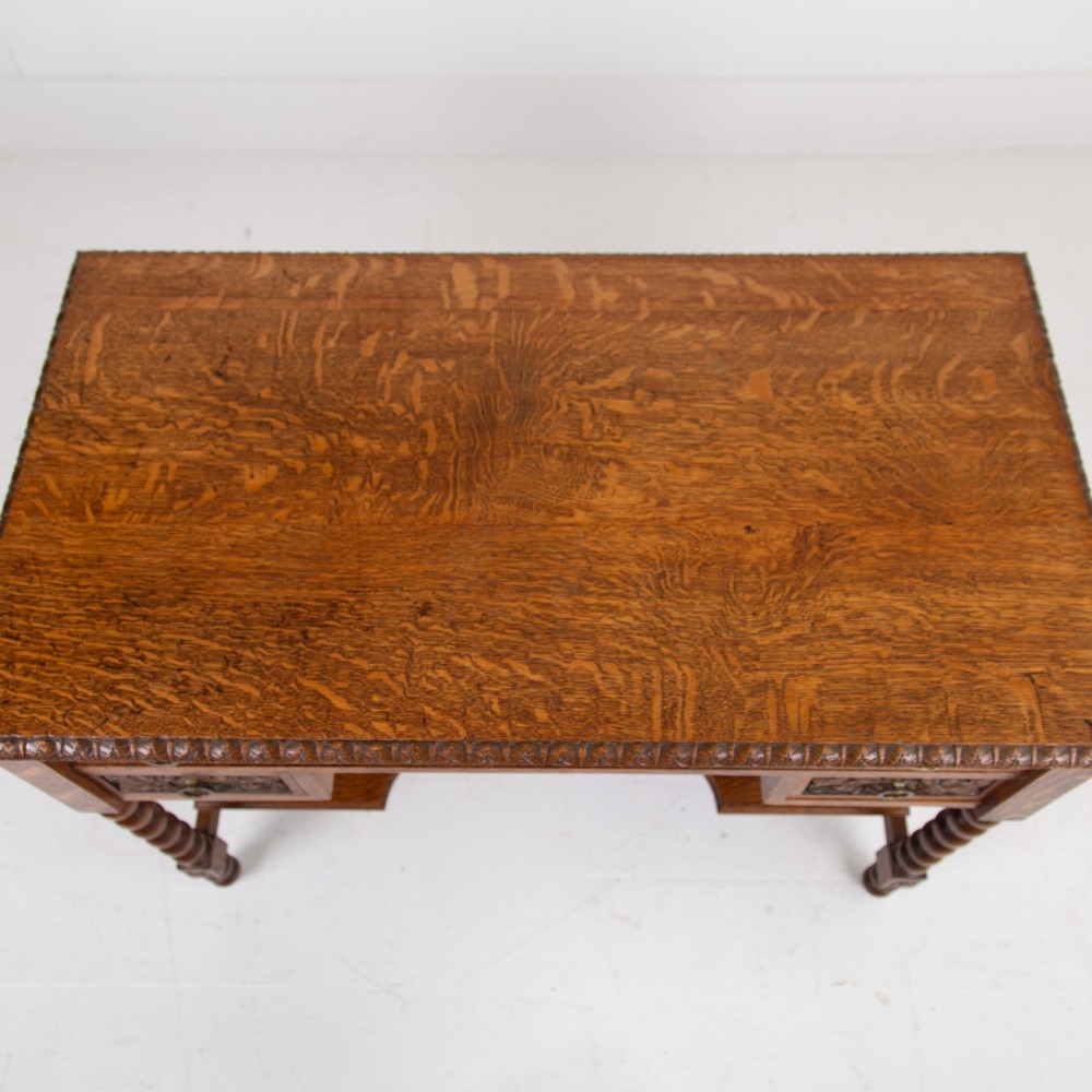 An oak writing desk featuring three carved foliage drawer fronts, bobbin-turned legs, brass drop handles, and a low under-tier shelf. View X