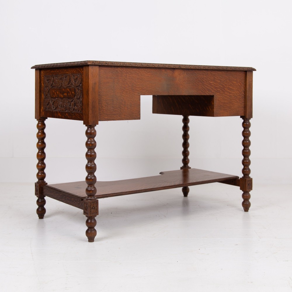 An oak writing desk featuring three carved foliage drawer fronts, bobbin-turned legs, brass drop handles, and a low under-tier shelf. View XI
