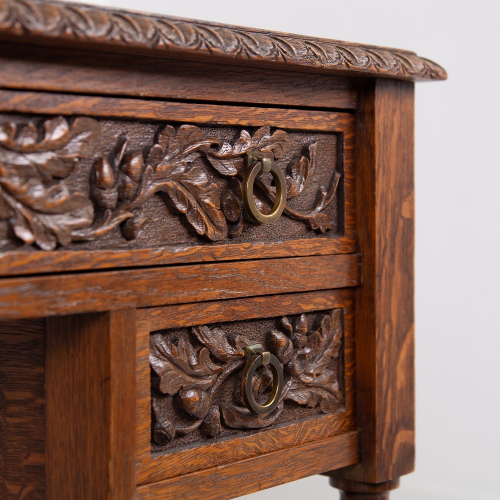 An oak writing desk featuring three carved foliage drawer fronts, bobbin-turned legs, brass drop handles, and a low under-tier shelf. View XII