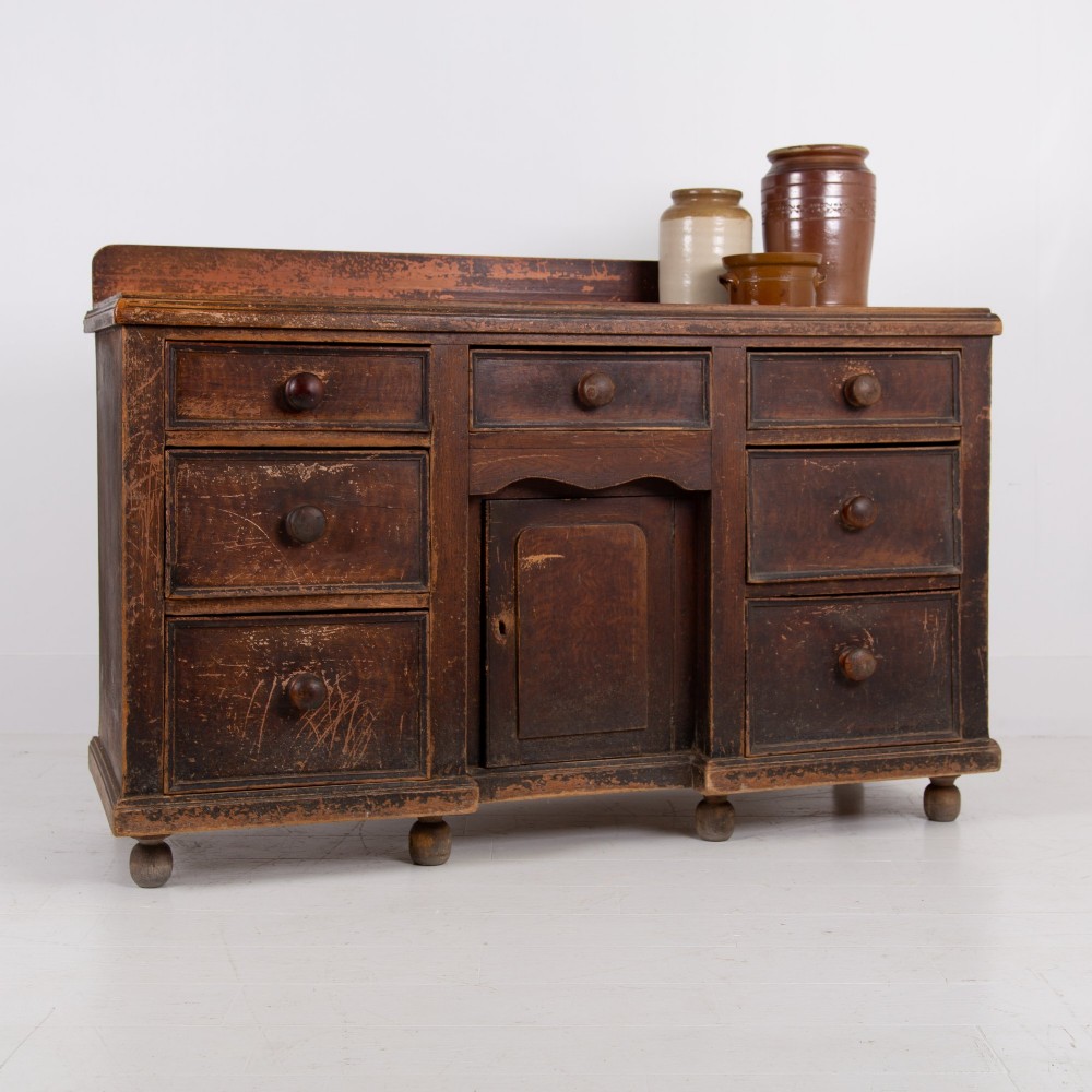 A 19th-century dark brown painted wooden kitchen dresser with seven drawers, a central cupboard, wooden knobs, and turned feet.