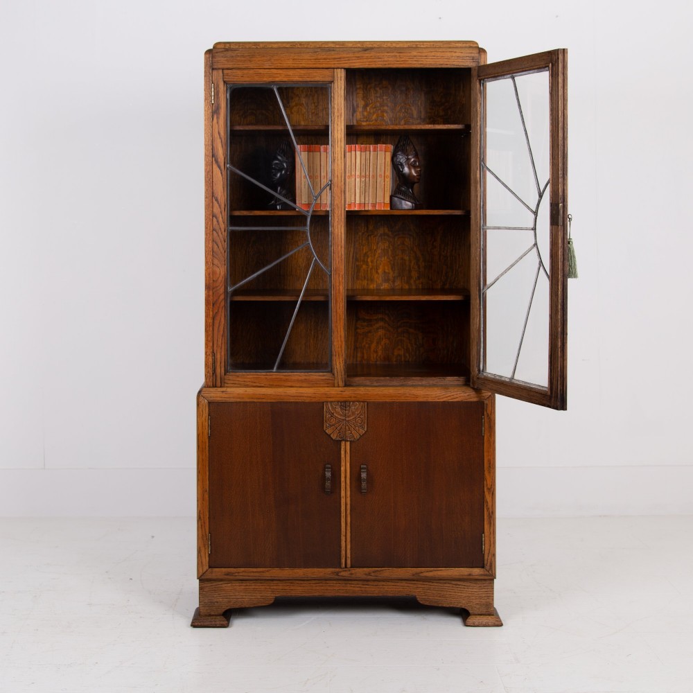 An Art Deco oak cabinet featuring two upper leaded glass doors with a geometric motif and a lower two-door wooden cupboard on stepped feet.