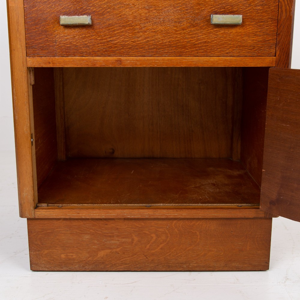 An Art Deco medium oak cabinet featuring two upper drawers, a lower cupboard door, and rectangular metal pulls on a plinth base. View X