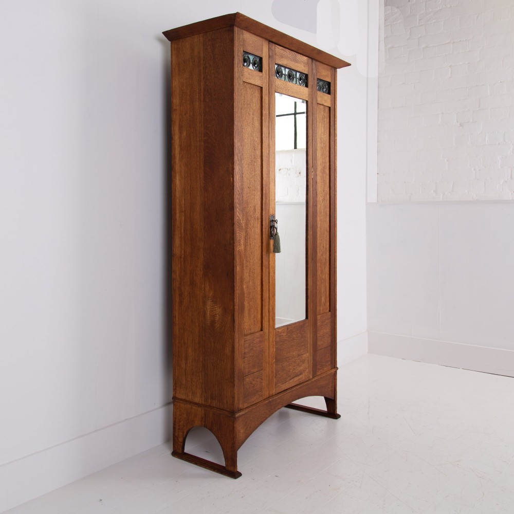An Arts and Crafts oak hall wardrobe featuring a central mirrored door, dark glass roundel inserts near the top, and an arched apron base. View II