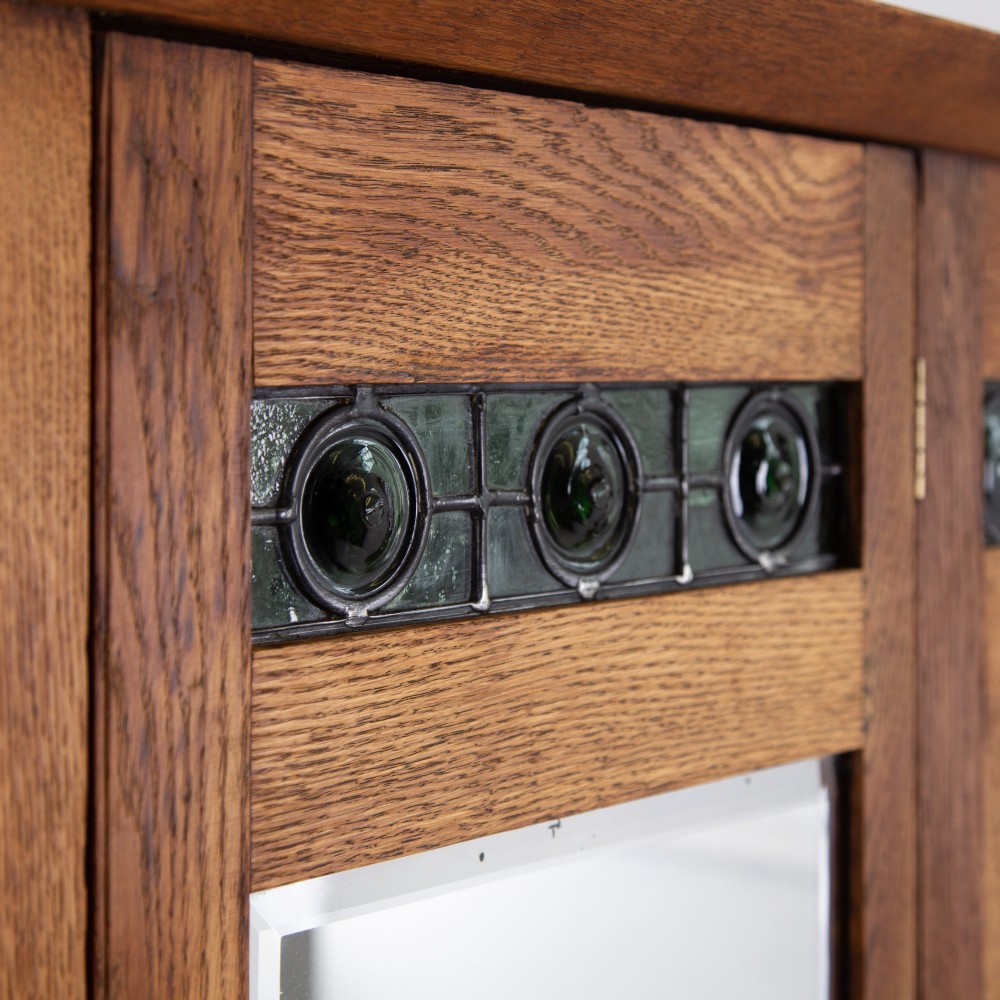 An Arts and Crafts oak hall wardrobe featuring a central mirrored door, dark glass roundel inserts near the top, and an arched apron base. View III