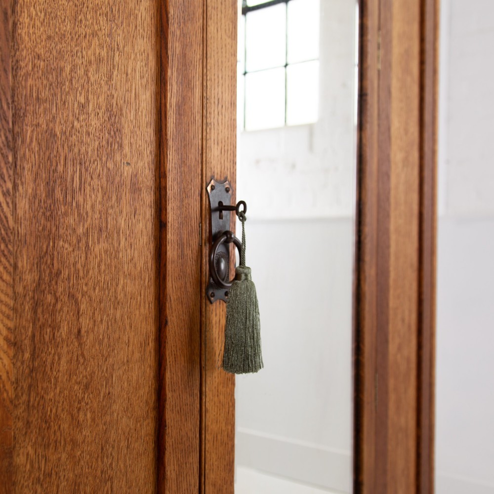 An Arts and Crafts oak hall wardrobe featuring a central mirrored door, dark glass roundel inserts near the top, and an arched apron base. View IV