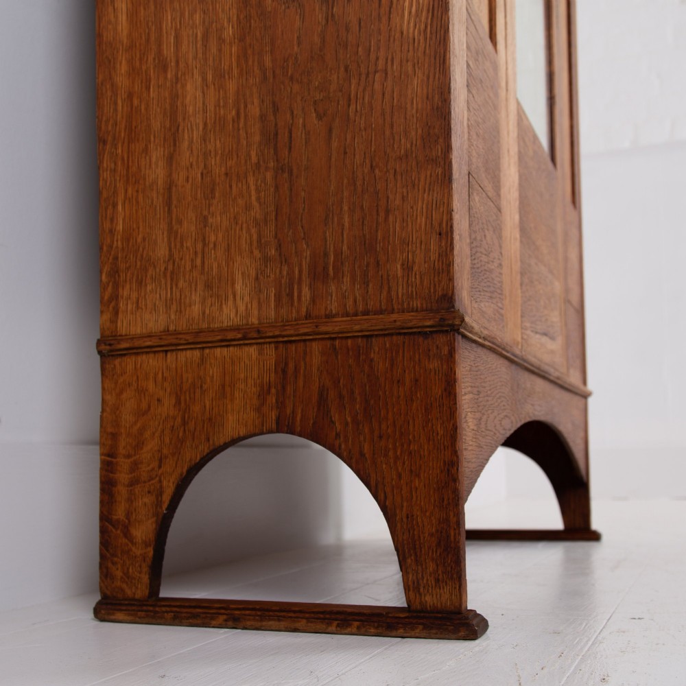 An Arts and Crafts oak hall wardrobe featuring a central mirrored door, dark glass roundel inserts near the top, and an arched apron base. View VI