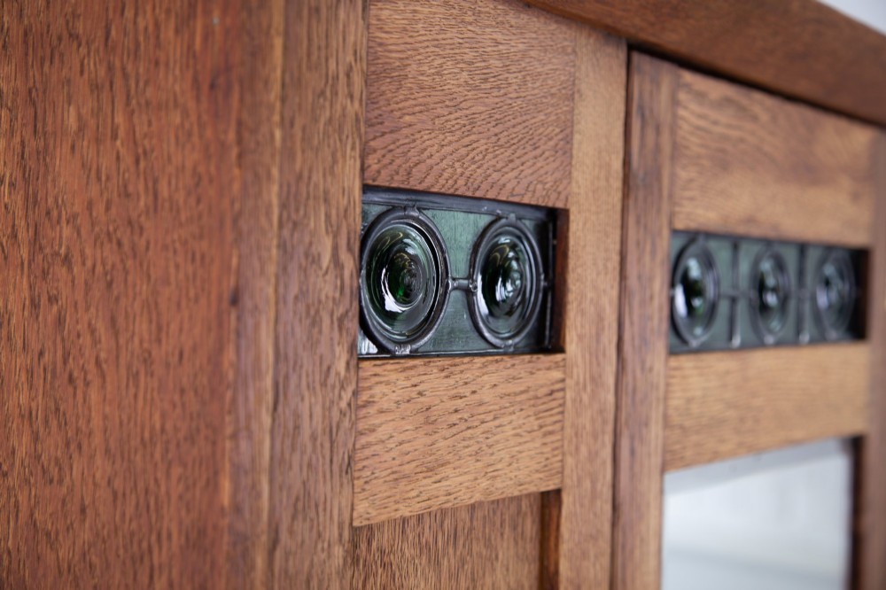 An Arts and Crafts oak hall wardrobe featuring a central mirrored door, dark glass roundel inserts near the top, and an arched apron base. View VII
