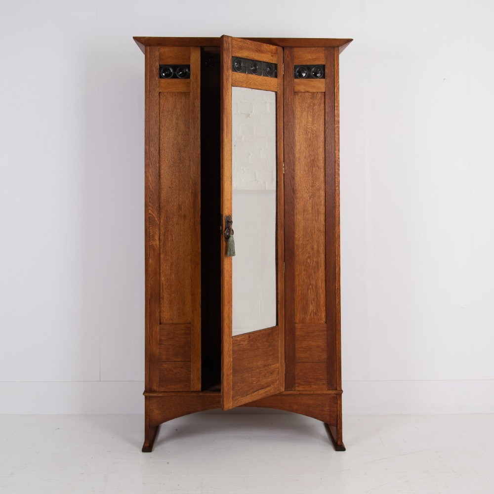 An Arts and Crafts oak hall wardrobe featuring a central mirrored door, dark glass roundel inserts near the top, and an arched apron base. View IX