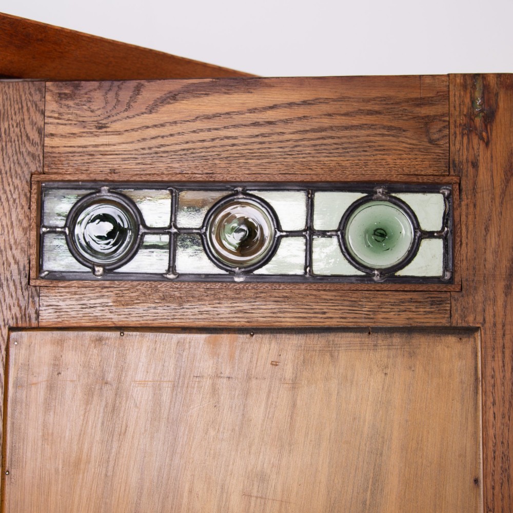 An Arts and Crafts oak hall wardrobe featuring a central mirrored door, dark glass roundel inserts near the top, and an arched apron base. View X